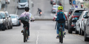 Zu sehen ist eine Straße. Auf dieser fahren zwei Kinder mit Rucksäcken Fahrrad. Sie sind nur von hinten zu sehen. Links und rechts der Straße parken Autos. Beide Kinder tragen einen Helm.