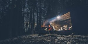 Eine Person wärmt sich am Feuer vor einer Waldhütte