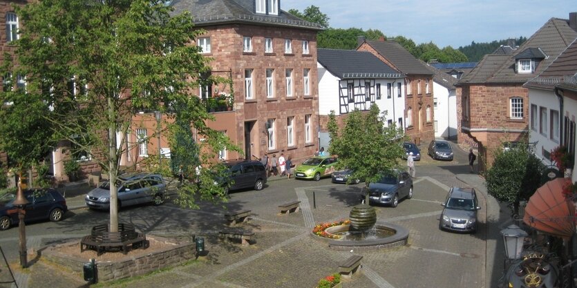 Ein Marktplatz. Links im Bild eine etwas erhöht stehende Linde, die von einer Bank umschlossen ist