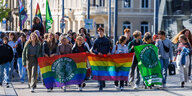 Zu sehen sind junge Demonstrierende auf der Straße die auf die Kamera zulaufen. Sie halten Regenbogenflaggen. Eine hat einen grünen runden Aufdruck auf dem Fridays for Future steht.