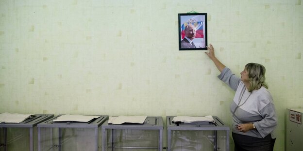 A woman hangs a picture of Vladimir Putin on the wall.  Below are stacks of ballot papers