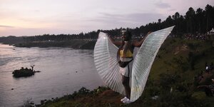 Eine Frau mit Flügeln steht an einem Gewässer, im Hintergrund ein Musikfestival