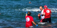 Drei Menschen, davon zwei in T-Shirts mit dem Symbol des Roten Halbmondes, stehen im flachen Küstengewässer, etwas weiter hinten schwimmt ein Mann.