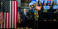 Biden steht neben einer US-amerikanischen Flagge und Menschen, die auf einer Bühne stehen und Schilder hochhalten, auf denen "Union Strong", "A better America" und "USA" steht