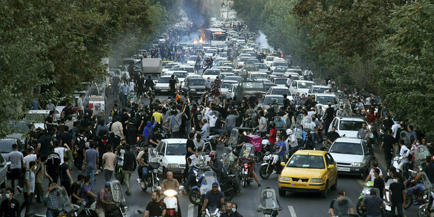 People and cars at protests in Tehran, in the background a fire and clouds of smoke