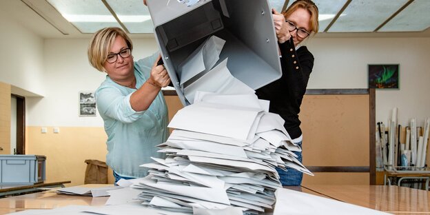 Two women tip a bin full of ballot papers onto a table