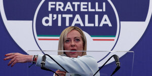 Georgia Meloni steht an einem Redepult vor einem Schriftzug ihrer Partei "Fratelli d'Italia" und zeigt mit dem linken Arm nach rechts.