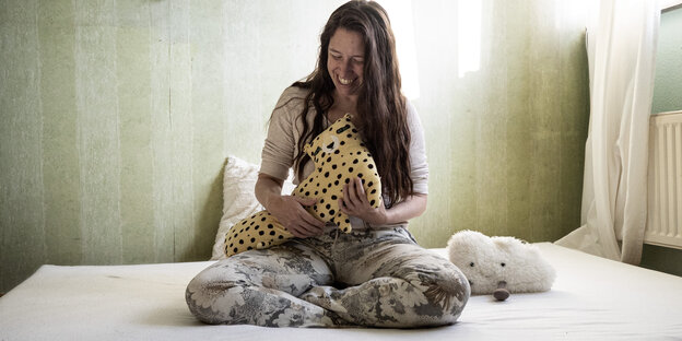 A woman sits on a bed and smiles, in her arms a plush leopard, next to her a cuddly cloud