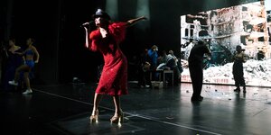 Schauspieler agieren vor einer Projektionswand, vorne singt eine Frau in einem roten Kleid