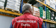Ein Mann mit roter Weste und Aufschrift "Hamburger Tafel" vor hohen Regalen mit Kisten drauf