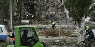 Ein vollkommen zerstörtes Gebäude im Bildhintergrund, ganz vorne ein Mann in einem grünen Bagger. Zudem ein Zivilist auf einem Fahhrad und zwei Männer in gelben Westen vor der Ruine.