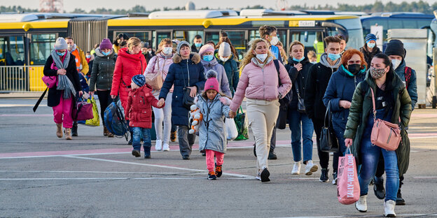 A group of women with small children is walking across a field, with yellow buses behind them
