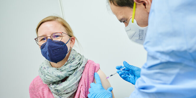 Zu sehen ist eine offensichtlich sitzende Frau. Im Bildausschnitt ist nur ihr halber Oberkörper sichtbar. Sie trägt eine dunkelblaue Maske und einen rosanen Pullover. Ihr rechter Pulloverärmel ist nach oben geschoben, ihr Arm ist frei. Eine Person in einem blauen medizinischen Kittel beugt sich zu der Frau herunter und setzt eine Spritze in ihren Oberarm. Die gebeugte Person trägt eine Maske und ist nur von der Seite und leicht verschwommen zu sehen.