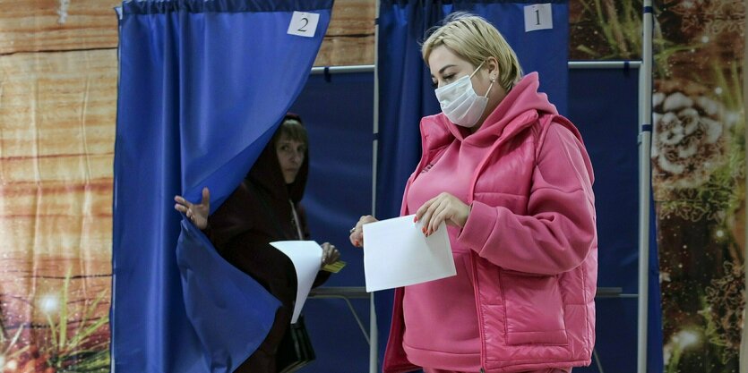 Zwei Personen in einem Raum, im Hintergrund einige Wahlkabinen.