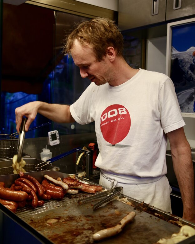 Ein Mann in weiße T-Shirt in einem Imbissstand, vor ihm auf dem Rost eine Wurst, daneben viele fertige Bratwürste, er nimmt gerade etwas mit einer Zange aus einem Fach