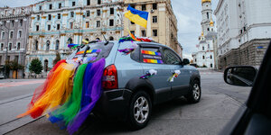 Ein Auto mit Regenbogen- und UKraineflagge vor zerstörten Wohnhäusern
