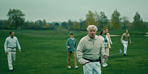 Eine Gruppe von Menschen mit Golfschlägern geht mit wachsamen Blicken über einen Rasen.