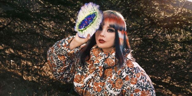 the Icelandic singer Björk sits in nature in front of leafy branches, has dyed her hair white-orange and black and holds a large artificial flower next to her head