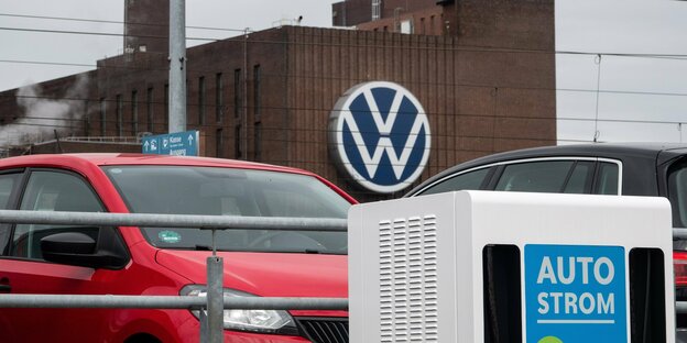 Eine Ladestation für Elektrofahrzeuge steht auf einem Parkplatz, im Hintergrund ist das Volkswagen Werk zu sehen.