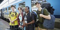 Die vier junge Menschen stehen vor einem blauen Zug an einem Bahnhof. Sie haben Rucksäcke auf.