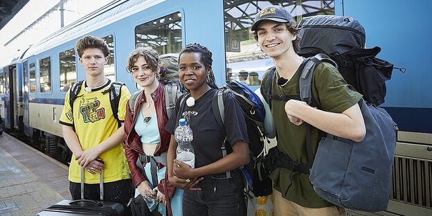 Die vier junge Menschen stehen vor einem blauen Zug an einem Bahnhof. Sie haben Rucksäcke auf.