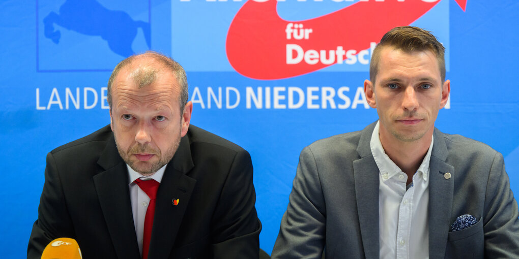 Zwei Männer sitzen an einem Tisch vor einer Wand mit dem Logo der AfD