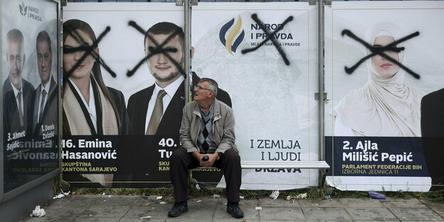 A man is sitting at a bus stop.  Behind him are election posters with paint smeared on their faces.