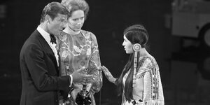 Ein schwarzweiß-Foto auf dem rechts die indigene Aktivistin Sacheen Littlefeather mit ihrer Hand die Oscar-Auszeichnung ablehnt, die die links-stehenden SchauspielerInnen Roger Moore und Liv Ullmann ihr überreichen wollen