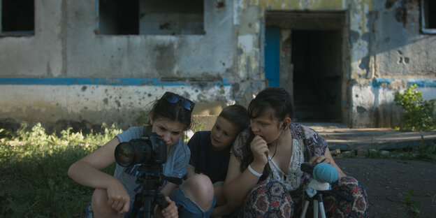 Drei Kinder sitzen vor einem leerstehenden Gebäude: Sie haben vor sich zwei Stative, links im Bild steuert ein älteres Mädchen eine Canon-Kamera, die Kinder blicken konzentriert auf das Display der Kamera