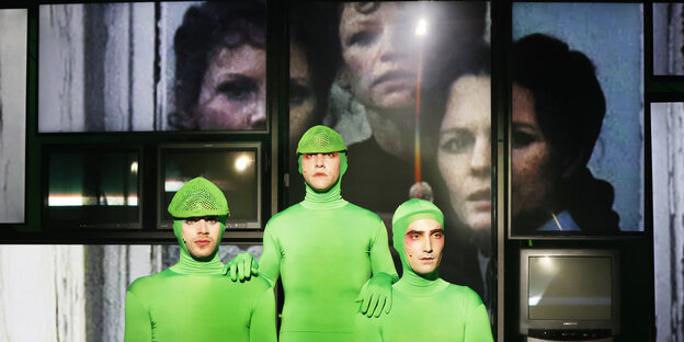 Drei Schauspieler in grünen Anzügen und Kappen stehen vor einer Projektion, in der man die Gesichter dreier Frauen erkennt