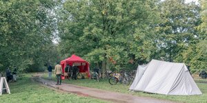Ein graues Zelt steht vor eiem überdachten Infostand in einem Park