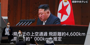 Kim Jong Un auf einem großen Bildschirm, wie er in Mirkofone spricht und vor der Nordkorea-Flagge sitzt