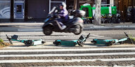 Drei E-Scooter liegen auf der Straße, im Hintergrund fährt eine Person mit dem Roller vorbei