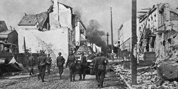 Schwarz-weiß Aufnahme eines teilweise zerstörten Dorfes, auf der Straße Soldaten und ein Panzerfahrzeug