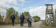Mit Helm, Schlagstock und Schild bewaffnet patroulieren drei Grenzpolizisten an einem Stacheldrahtzaun mit Wachturm