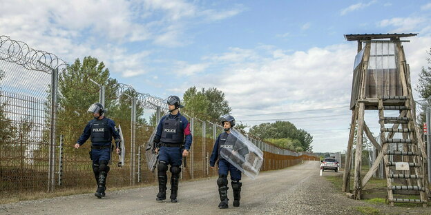 Armed with helmets, batons and shields, three border police officers patrol a barbed wire fence with a watchtower
