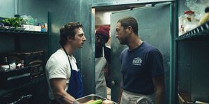 Zwei Männer in Kochschürzen stehen sich in einem Vorratsraum gegenüber, ein Dritter Mann schaut durch die Tür