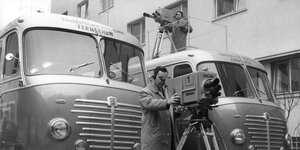 Ein Schwarz-Weiß-Foto. Ein Mann in Mantel steht vor zwei Bussen. Er schaut in eine Kamera. Auf dem Dach von einem der Busse steht noch ein Mann mit Kamera.