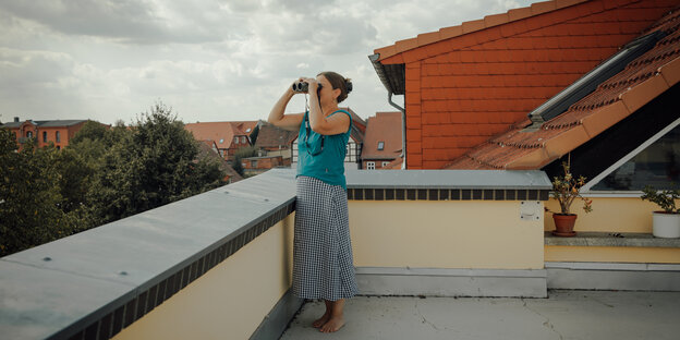 Eine Person steht mit Fernglas auf einer Dachterasse.