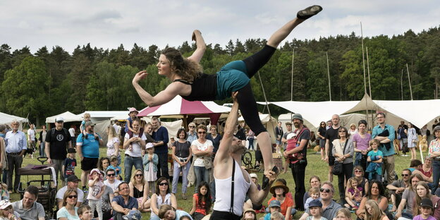 Bei einer Zirkus-Performance mit Publikum auf einer Wiese hält ein Mann eine Frau hoch