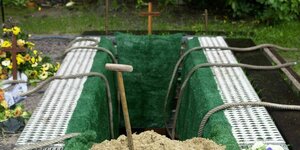 Offenes Grab mit grünem Kunstrasen und einem Sandhaufen davor, in dem eine Schaufel steckt