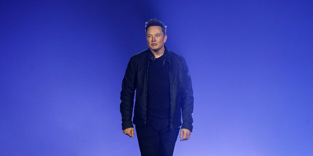 Elon Musk vor einer blauen Wand. Er hat kurze dunkle Haare trägt schwarze Hose und Shirt und eine schwarze Lederjacke. Er schaut grimmig.