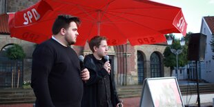 Der junge SPD-Landtagskandidat Besian Krasniq steht neben dem SPD-Generalsekretär Kevin Kühnert unter einem roten SPD-Schirm auf dem Marktplatz von Quakenbrück.