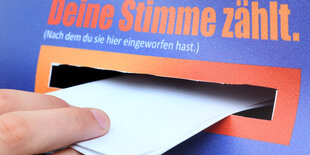 Eine Kinderhand schiebt einen Stimmzettel in einen blauen Karton mit der Aufschrift "Deine Stimme zählt. (Nachdem du sie hier eingeworfen hast)."