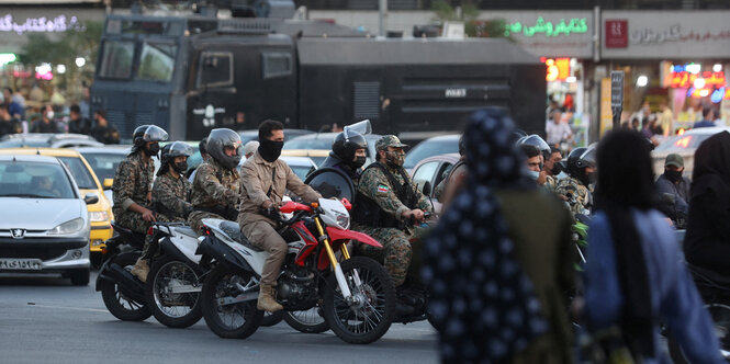 Männer in Militärkleidung auf Motorrädern. Auf der Straße schauen ihnen Frauen aus der Ferne zu.