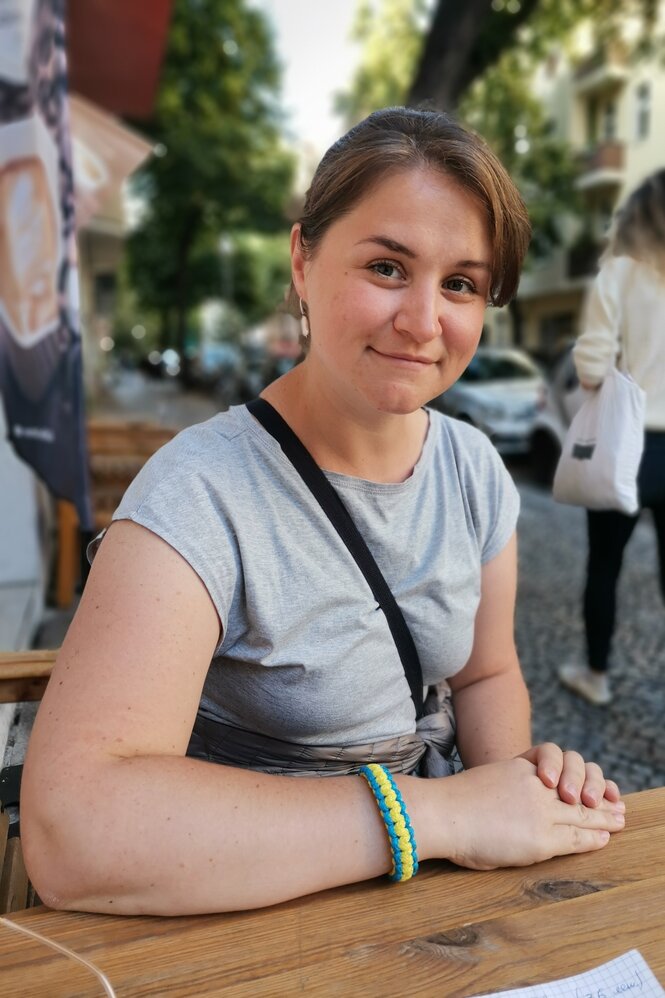 Eine junge Frau mit grauem shirt schaut in die Kamera und hat ihre Hände auf einem Tisch afugelegt