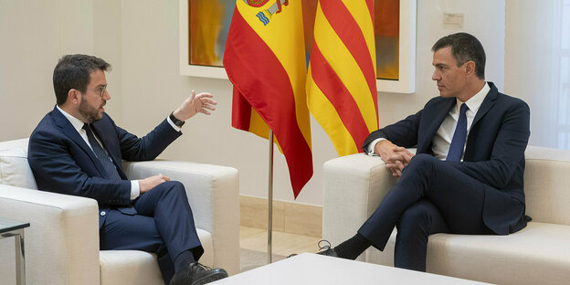 Aragonès und Sánchez sitzen in einer weißen Polsterlandschaft und unterhalten sich. Im Hintergrund hängen die spanische und die katalonische Fahne