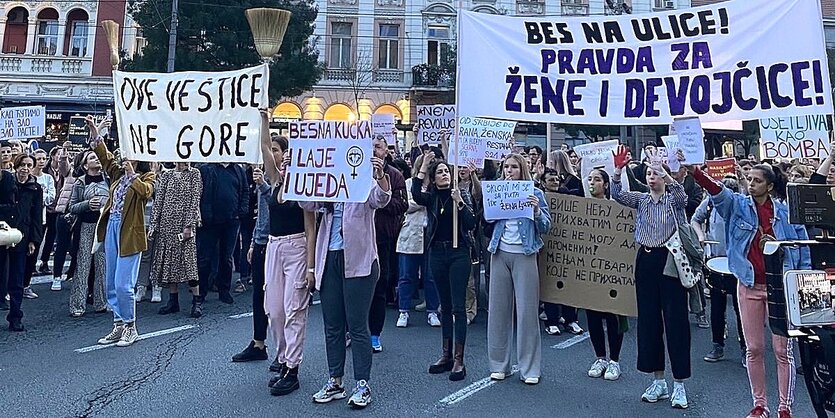 Mehrere Demonstrantinnen halten auf einer Straße Schilder und Banner mit serbischer Beschriftung in die Höhe