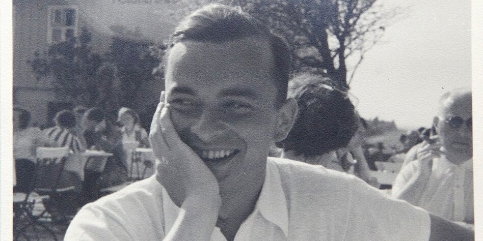 Ein junger Mann sitzt in einem Biergarten und lächelt mit aufgestützter Hand