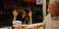 Filmstill: Eine junge Frau legt ihre Hände auf eine Schaufensterscheibe und blickt dadurch einen Mann, der in einem Diner beim Essen sitzt